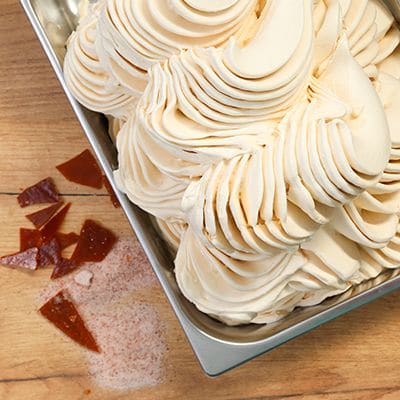 Jasne lody karmelowe ułożone falami w kuwecie, obok kawałki karmelu i różowa sól