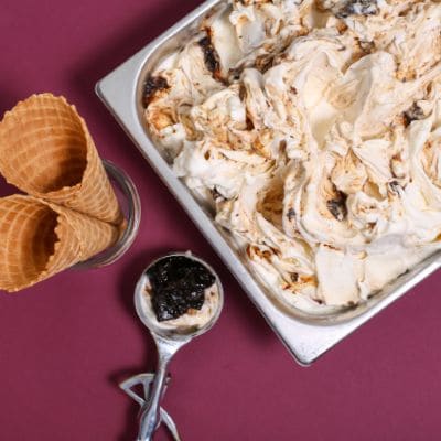 Jasne lody z ciemnymi wstęgami owoców w kuwecie, obok rożki i gałkownica na bordowym tle